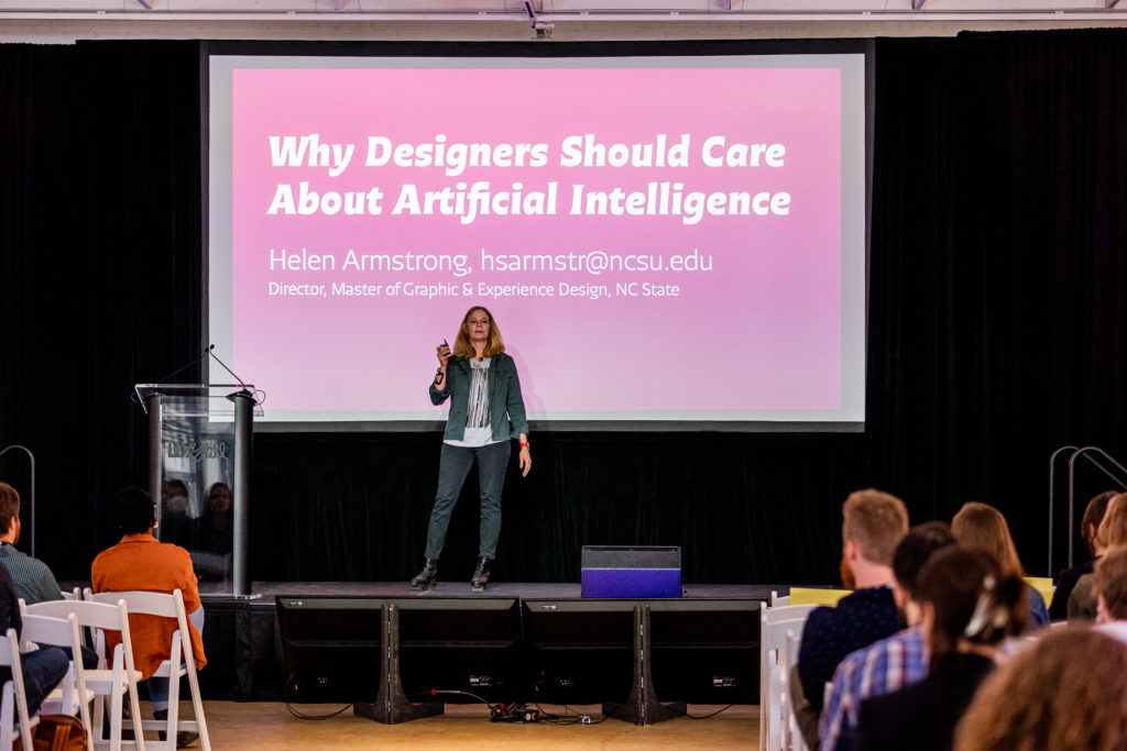 White women with blonde hair in jeans and a green jacket on stage in front of large screen. Screen reads: Why Designers Should Care About Artificial Intelligence.