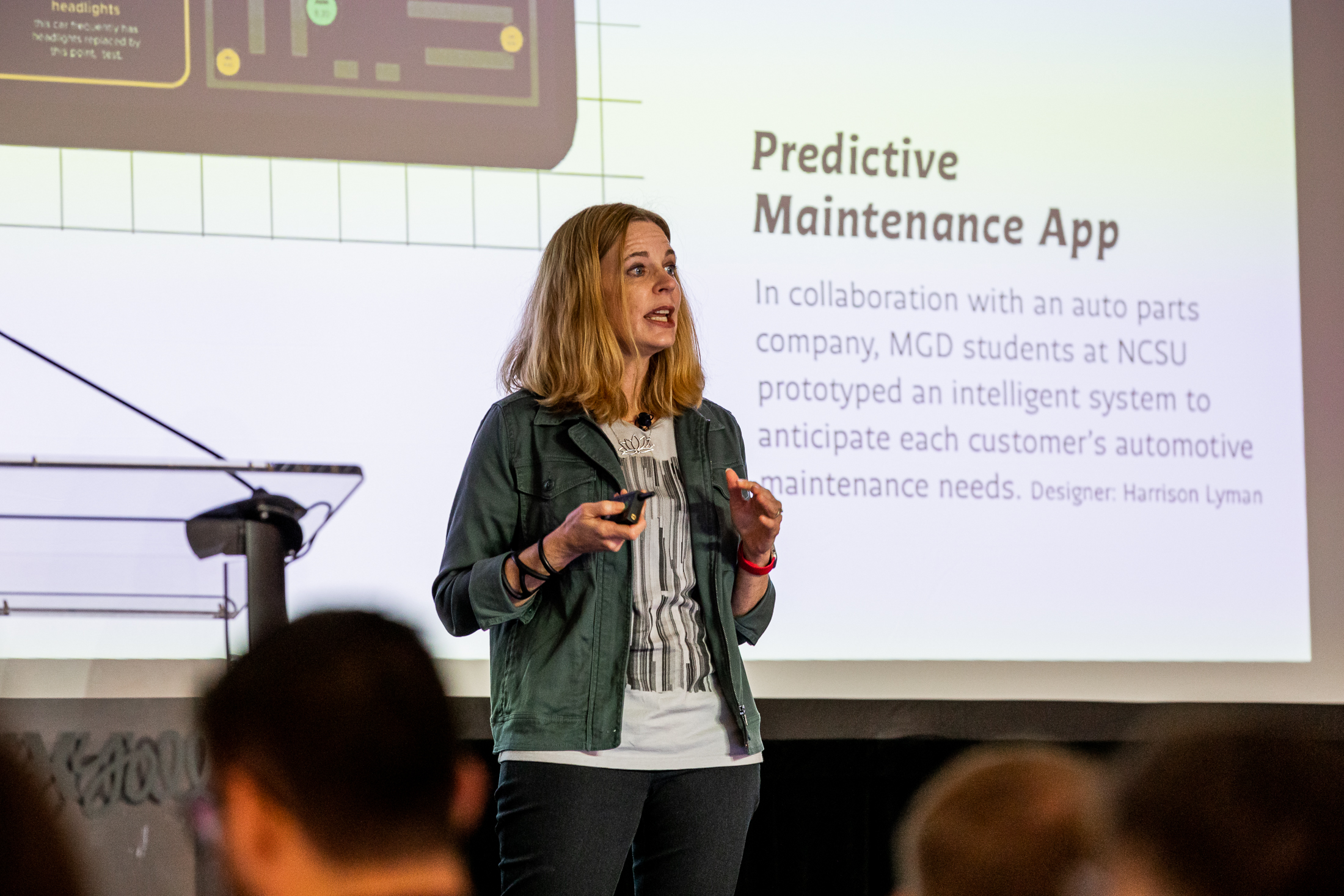 white women with blonde hair on stage. She is speaking in front of a large screen. Screen reads: "Predictive Maintenance App"